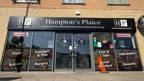 Ben Schofield/BBC The exterior of Hampton's Plaice fish and chip shop. A set of double doors is in the centre of image, with two windows on either side of the doors. The windows bear words including "pizzas", "sausages & pies", and "desserts". Under "Hampton's Plaice" is written: "Serving our local community with quality fish and chips". The shop front is set into a brick-built building and above the shop the very bottom of three windows are just shown. 