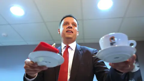 PA Media Anas Sarwar holding a cup of coffee in his left hand and a saucer with a red napkin in his right hand. He is smiling and wearing a dark suit, white shirt and red tie