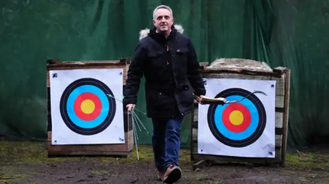 PA Media Alex Cole-Hamilton walks towards the camera, holding a bow in his left hand and arrows in his right hand. There are two large targets behind him and he is wearing a dark blue jacket with a furry hood and blue jeans.