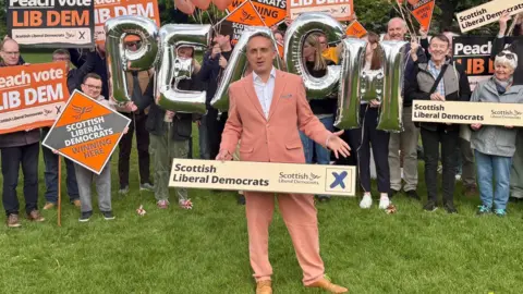 PA Media Alex Cole-Hamilton holding a Scottish Liberal Democrats banner wearing a peach suit and standing in front of supporters holding placards