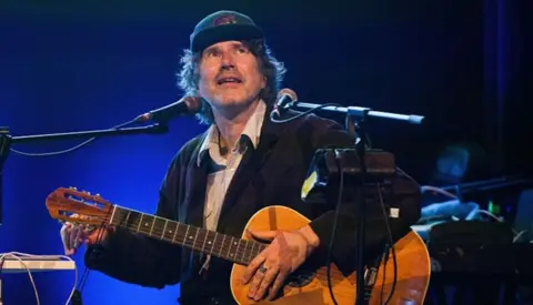 Getty Images Gruff Rhys, on stage with an acoustic guitar, dressed in a cap and dark jacket, smiling at the crowd.
