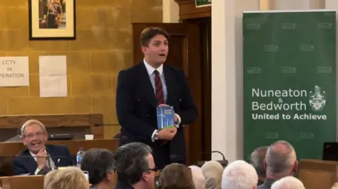 George Finch holds a Ladybird Book on Climate Change in a meeting of Nuneaton and Bedworth Borough Council. He is wearing a blue suit and striped red and blue tie and is standing. There are numerous councillors sitting facing Finch in the room with their backs to the camera. One man to the left is facing the camera and is sat next to Finch. He has grey hair and is smiling.