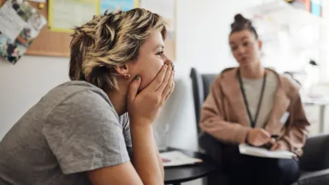 Getty Images Stock image shows a young person with short blonde hair with their hands over their mouth looking pensively away from camera while a counsellor in the background speaks and writes on a piece of paper.