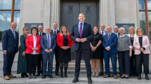 Welsh government The first minister and cabinet ministers stand on steps in front of a grey building with a brown door.