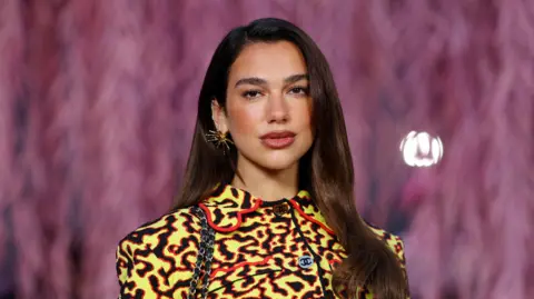 Reuters Dua Lipa, a young woman with long brown hair and a yellow and black patterned top, poses for the camera at a Chanel fashion show in Paris