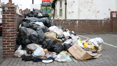 EPA/Shutterstock A large pile of rubbish in a small private parking area on a suburban street. Black and white bin bags have spilled open, with soiled flattened carboard and other detritus on the floor. Homes and for sale signs can be seen behind.