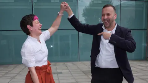 PA Media Zoë Garbett, left, seen from side view, laughs, smiles and does a high hand-shake with Jack Zack Polanski as they celebrate her mayoral win. She has short hair which is dark apart from a pink fringe and wears a white blouse and red skirt. He wears dark suit and white shirt.