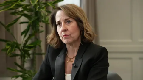 Liz Kendall, wearing a black suit jacket, gold necklace and white top
