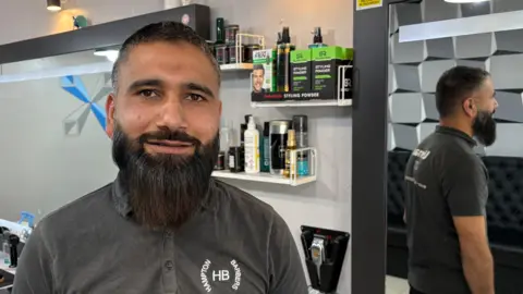 Ben Schofield/BBC Muhammad Usman, standing inside a barber shop, looking down the camera and pictured on the left of the frame. He has dark brown eyes, a relatively long straight beard and short hair. He is wearing a grey, collared t-shirt, on which the logo of "Hampton Barbers" can be seen. Hair care products are on shelves mounted on the wall behind Muhammad. The shelves are next to a large mirror, in which Muhammad is reflected.
