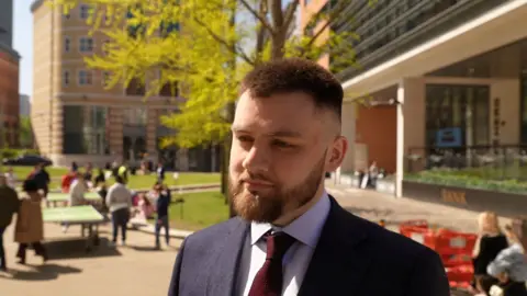 A man stands in a city square. He is wearing a navy jacket, grey shirt and maroon tie. He has spiky brown hair and manicured beard. He is looking to the right of the camera at something. Passers-by can be seen in the square behind him, the Bank pub, a Caffe Nero and grassy areas. It is a sunny day.