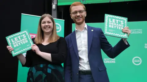 PA Media Gillian Mackay and Ross Greer holding up copies of the Scottish Green manifesto