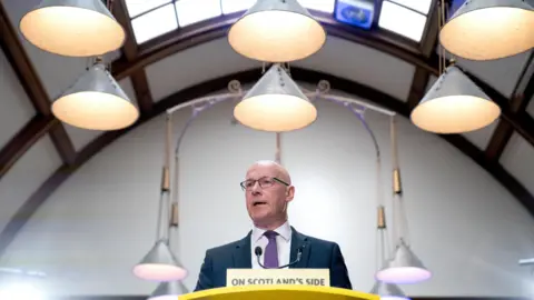 PA Media John Swinney, who is bald and wearing glasses, is standing behind a lectern in a domed room with lights hanging above his head. He is wearing a blue or grey suit, white shirt and purple tie. There is a sign in front of him that says On Scotland's Side 