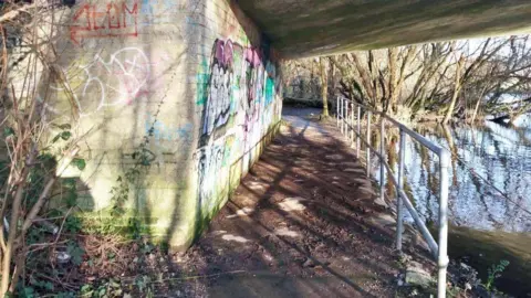 CPS An underpass beneath a bridge next to a river. The wall is covered in graffiti and there is a metal railing along the path.

