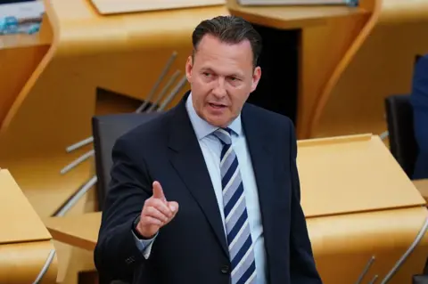 PA Media Scottish Conservative leader Russell Findally wearing formal business attire stands inside a modern parliamentary or legislative chamber with light wood desks arranged in rows. The individual is wearing a dark suit jacket, a light blue shirt, and a striped tie. One hand is raised with the index finger extended in a pointing gesture, suggesting emphasis during speech or debate. The background shows empty desks and chairs, softly lit with warm tones, indicating an indoor government or assembly setting.