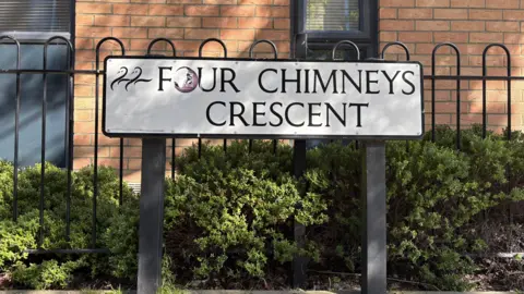 Ben Schofield/BBC A road sign bearing the street name "Four Chimneys Crescent". An outline of two swans is in the top left hand corner of the sign. The sign is against a row of black railings, behind which is growing a low, green hedge. Further in the background is a brick wall.