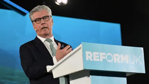 PA Media Malcolm Offord standing on stage behind a lectern with Reform UK written on it. He has grey hair and glasses and is wearing a dark suit, white shirt and green tie