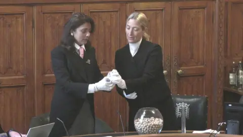 House of Commons A still showing Nusrat Ghani, the senior deputy speaker, being handed a ballot ball by a  parliamentary clerk. They are standing in a wood panelled room in the Commons, between green leather chairs and a desk. On the desk sits a clear glass goldfish bowl, which is filled with small, white ballot balls. The two women wear formal black dress coats and white gloves. Ghani wears her black hair long and loose, whereas the clerk has her long blonde hair tied back in a ponytail. 