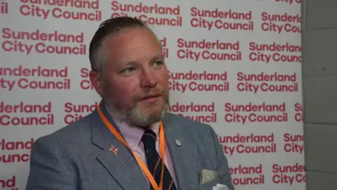 Reform UK councillor Paul Donaghy. He is wearing a grey suit with a Union Jack flag on the lapel. He has brown hair and a beard, both of which are graying.