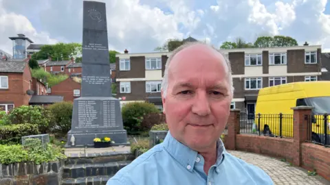 Tony Doherty is wearing a blue shoirt and is standing in front of the Bloody Sunday memorial in Derry