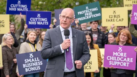 PA Media John Swinney standing in front of a group of MSPs and supporters holding signs in support of the SNP.