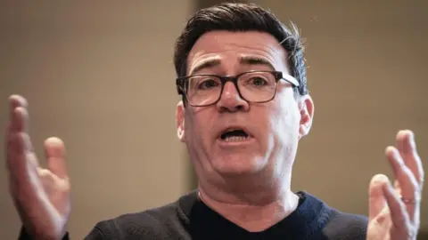PA Media Andy Burnham is wearing a black jumper and black glasses. He is speaking with his hands raised and held apart.