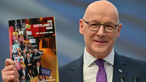 Getty Images John Swinney, in dark blue suit with white shirt and purple tie, is holding up the SNP's election manifesto, which is called On Scotland's Side. He is smiling and wearing spectacles and a saltire badge on his left lapel. 