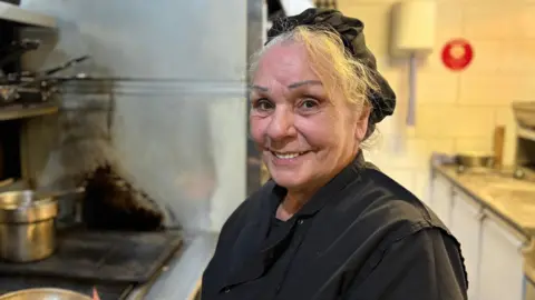 Ben Schofield/BBC Tina Defalco, smiling and looking straight down the camera. She is standing in a kitchen, next to a stove. She is wearing a black cotton chef's coat and a black hairnet, under which her grey, wavy hair is escaping. Behind her on the left is a stainless steel piece of equipment, while on the right is a work surface and a white tiled wall, on which a paper towel dispenser is mounted.